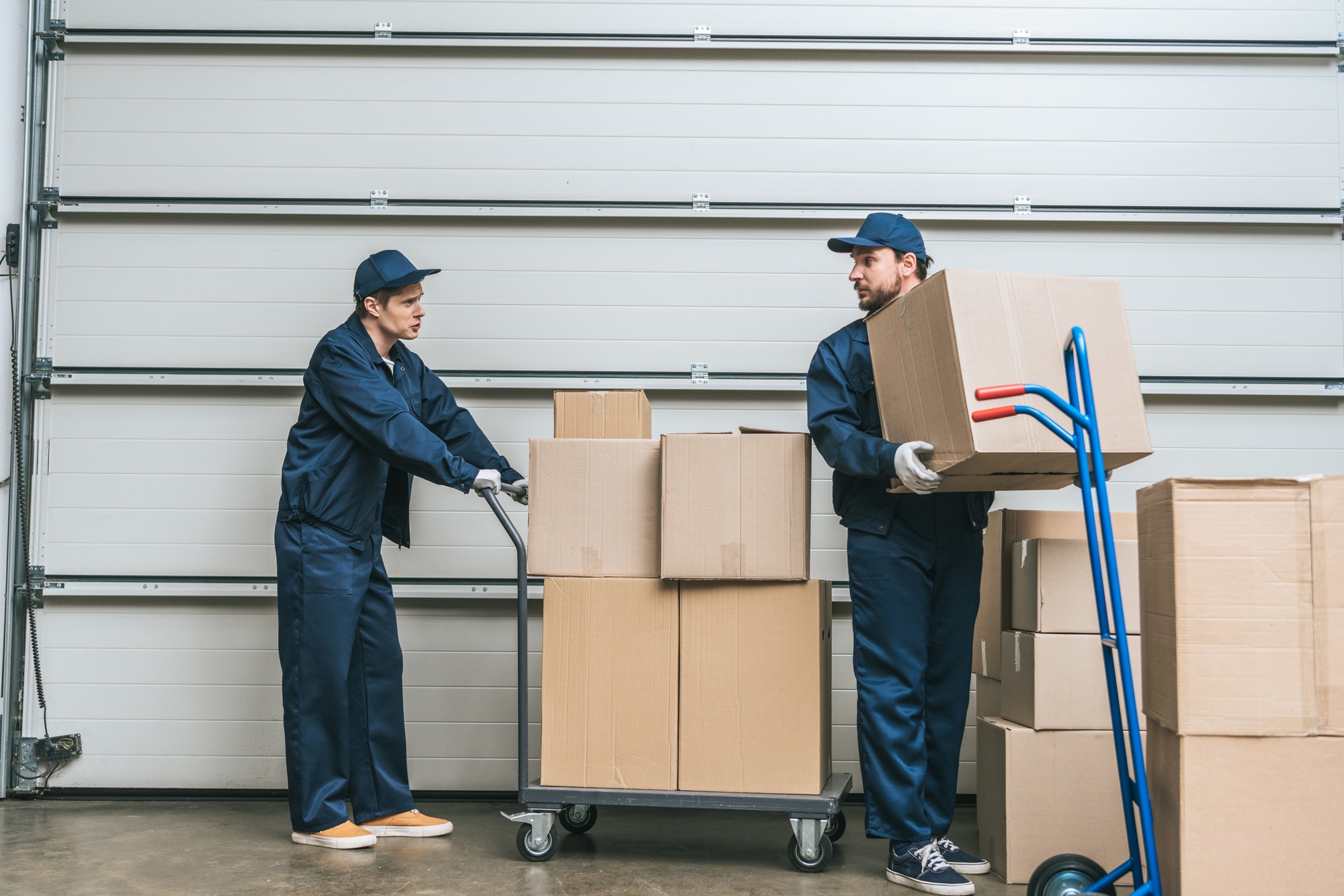 two movers in uniform looking at each other while transporting cardboard boxes with hand trucks in